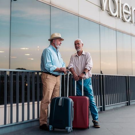 Zwei ältere Männer stehen mit ihren Rollkoffern am Flughafen und warten.